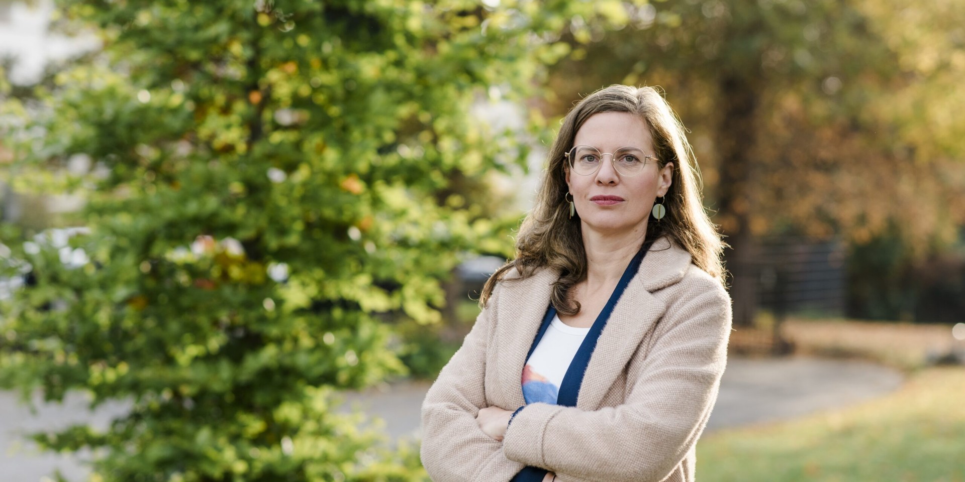 Frau mit Brille und beigem Mantel steht mit verschränkten Armen vor einem Baum.